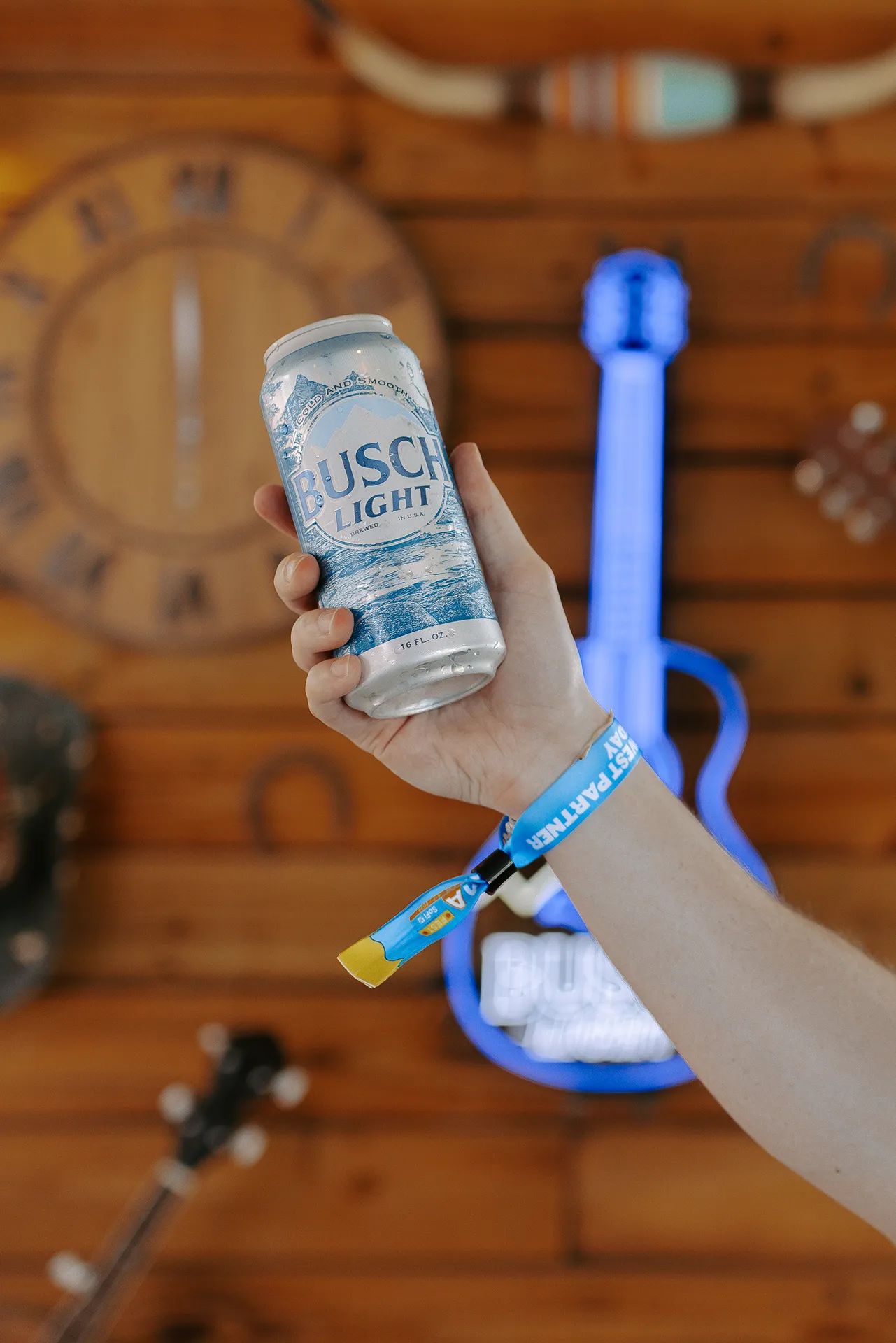 Hand holding a cold Busch Light can with a festival wristband against a timber wall